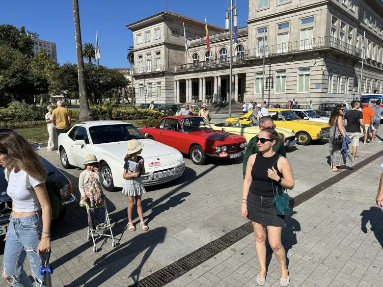 A white Porsche 944 Turbo, red Lancia Fulvia Sport, and yellow MGB roadster at a car show in Pontevedra, with Andrea in the foreground.