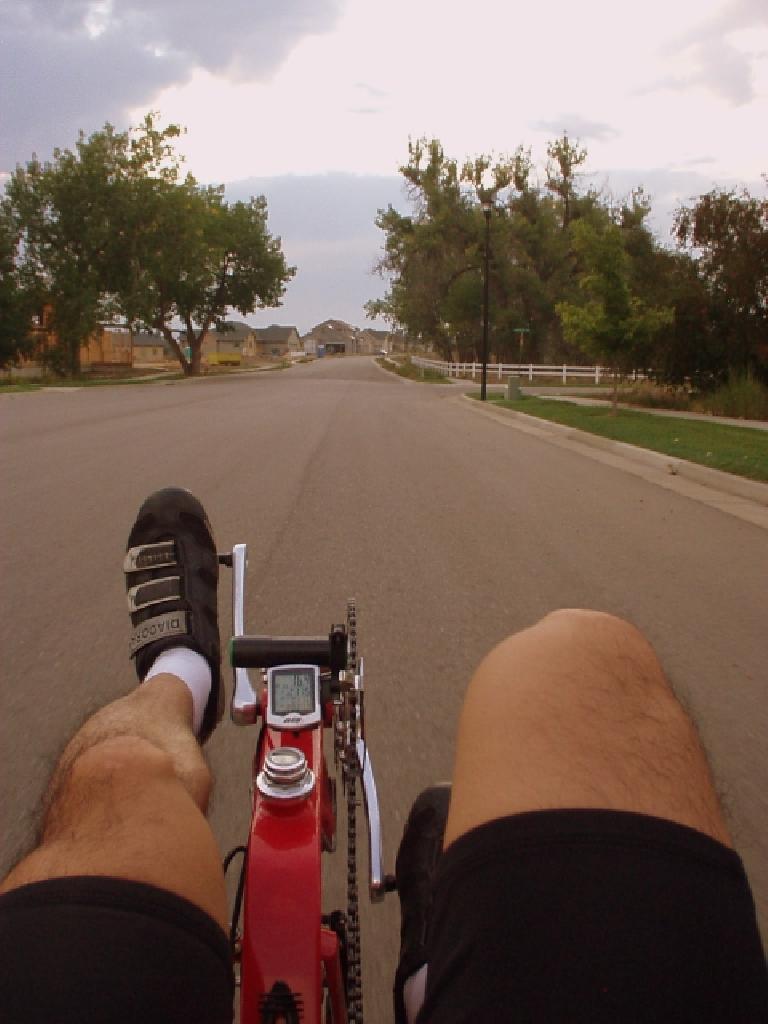 view of legs pedaling a red Reynolds Wishbone recumbent down the street towards some trees with speedometer reading 16.4 MPH