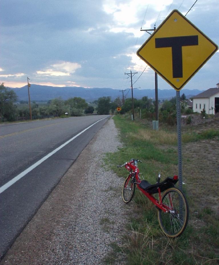 red Reynolds Wishbone recumbent leaned against a signpost with a yellow T-intersection sign, with Front Range foothills in the background