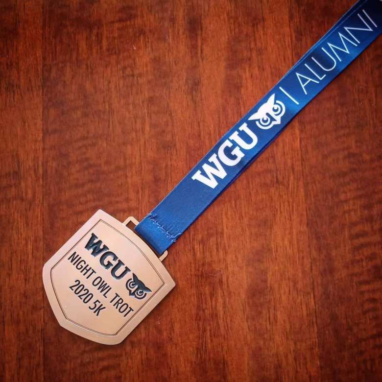 gold-colored medal that says WGU Night Owl Trot 2020 5K attached to blue lanyard that says WGU Alumni